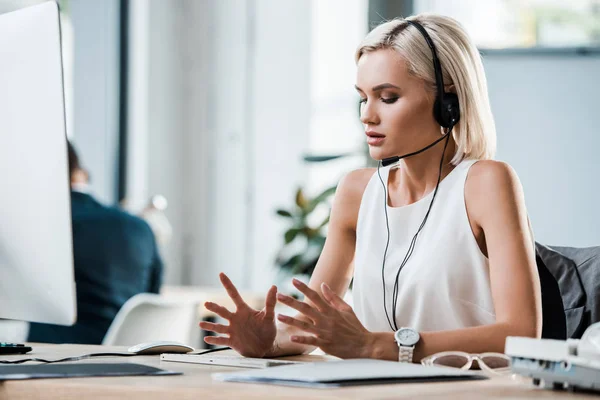 Attractive blonde woman in headset gesturing near computer monitor in office — Stock Photo