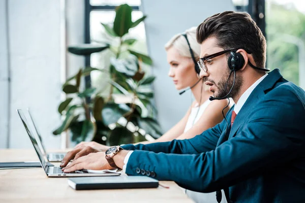Selective focus of handsome man in headset typing on laptop while working near colleague — Stock Photo