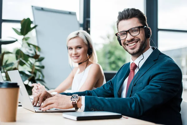 Selektiver Fokus eines fröhlichen Mannes im Headset, der auf dem Laptop tippt, während er neben einem blonden Kollegen sitzt — Stockfoto