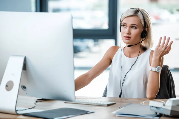 Selektiver Fokus der schönen blonden Frau im Headset, die im Büro in der Nähe des Computermonitors arbeitet — Stockfoto