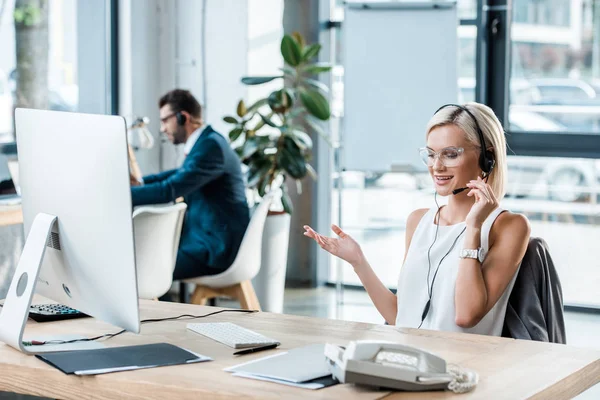 Enfoque selectivo de mujer rubia alegre en el gesto de auriculares mientras habla cerca de compañero de trabajo - foto de stock