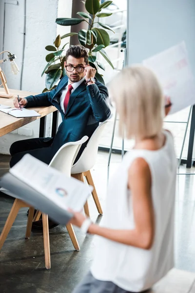 Selektiver Fokus eines gutaussehenden Geschäftsmannes, der eine Brille anfasst und einen blonden Kollegen mit Dokumenten ansieht — Stockfoto