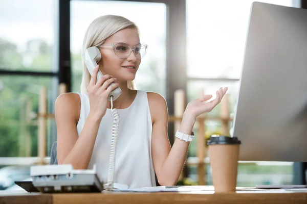 Selektive Fokussierung der blonden Geschäftsfrau, die beim Gesten und Sprechen auf dem Retro-Handy in der Nähe von Pappbechern lächelt — Stockfoto