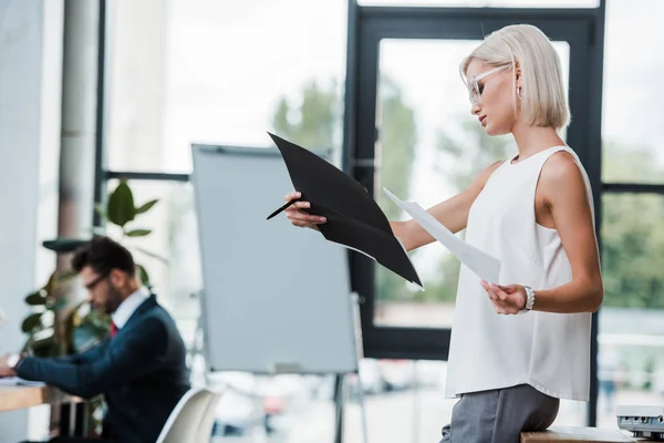 Messa a fuoco selettiva della donna bionda in occhiali tenendo penna cartella vicino al collega in ufficio moderno — Foto stock