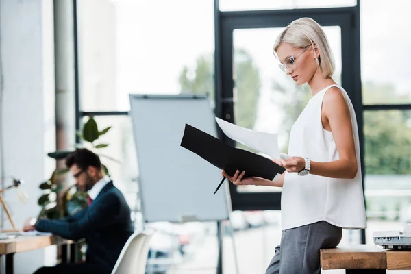 Selective focus of blonde businesswoman in glasses holding folder and documents near coworker in modern office — Stock Photo