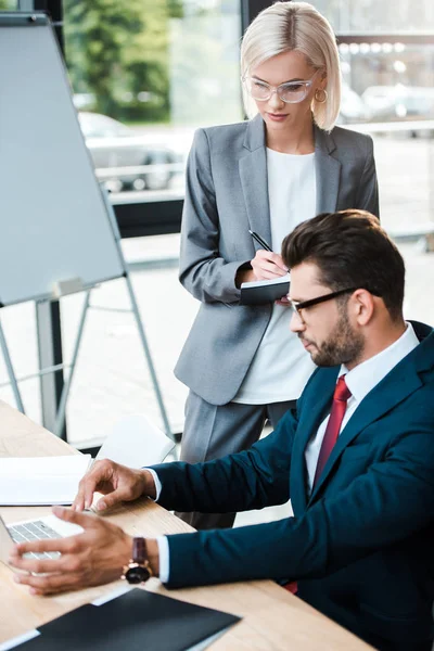 Messa a fuoco selettiva della donna attraente che scrive nel taccuino vicino all'uomo bello usando il computer portatile in ufficio — Foto stock