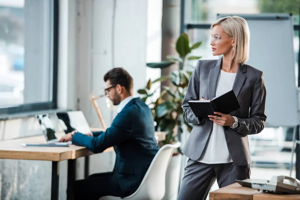 Selektiver Fokus der blonden Geschäftsfrau, die ihr Notizbuch in der Nähe ihres Büromitarbeiters hält — Stockfoto