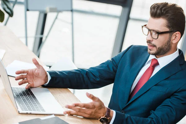 Guapo hombre de negocios en gafas de gestos mientras mira el ordenador portátil - foto de stock