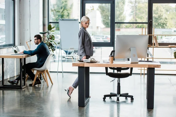 Attractive businessman in glasses looking at table while walking in office near coworker — Stock Photo