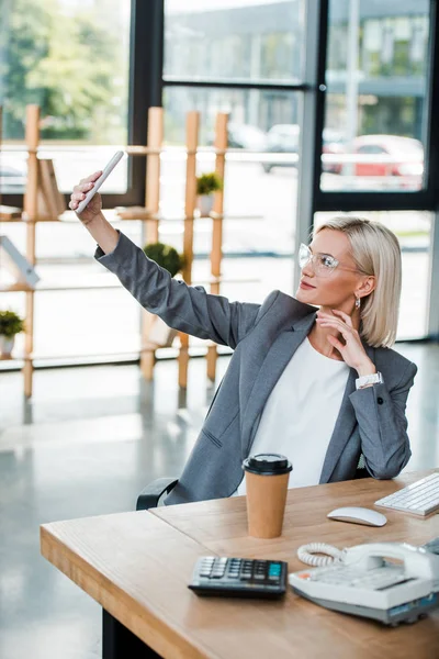 Вибірковий фокус бізнес-леді в окулярах приймає селфі в сучасному офісі — стокове фото