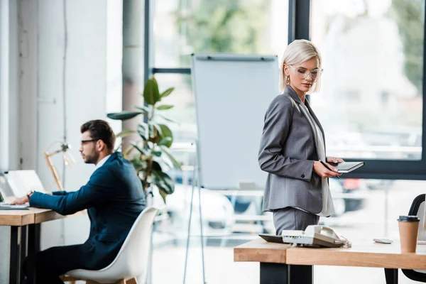 Enfoque selectivo de atractiva mujer de negocios rubia sosteniendo portátil oreja compañero de trabajo - foto de stock