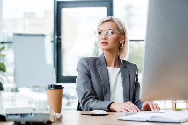 Selektiver Fokus der schönen Geschäftsfrau in Brille, die im modernen Büro in der Nähe von Pappbechern arbeitet — Stockfoto