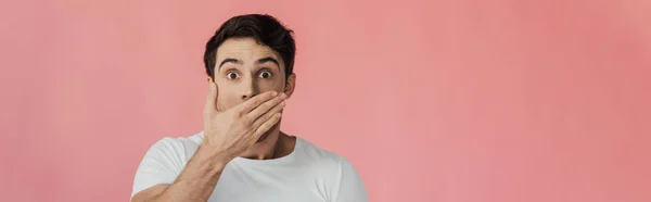 Panoramaaufnahme eines verängstigten jungen Mannes, der den Mund mit der Hand bedeckt und isoliert auf rosa Kamera schaut — Stockfoto