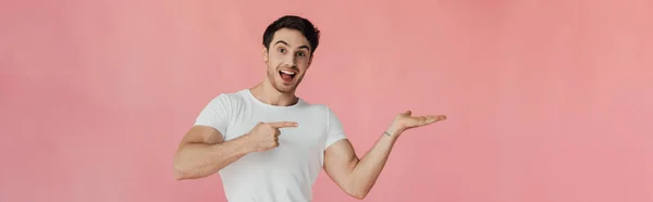 Panoramaaufnahme eines lächelnden jungen Mannes im weißen T-Shirt, der mit dem Finger isoliert auf rosa zeigt — Stockfoto