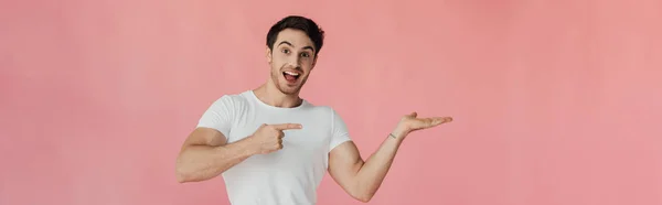 Tiro panorâmico de jovem sorridente em t-shirt branca apontando com o dedo isolado em rosa — Fotografia de Stock