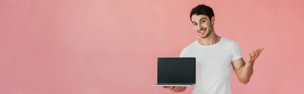 Panoramaaufnahme eines lächelnden jungen Mannes im weißen T-Shirt, der einen Laptop mit leerem Bildschirm zeigt, isoliert auf rosa — Stockfoto