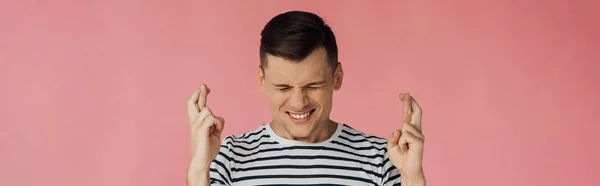 Panoramic shot of front view of smiling man in striped t-shirt with closed eyes and crossed fingers isolated on pink — Stock Photo