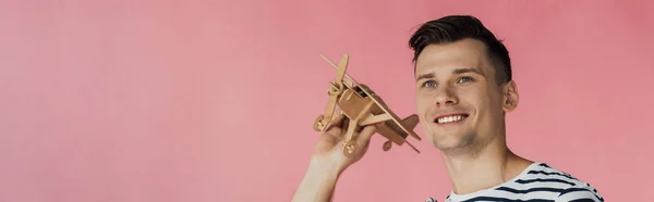 Tiro panorâmico de sorrir jovem segurando avião de brinquedo de madeira e olhando para longe isolado em rosa — Fotografia de Stock