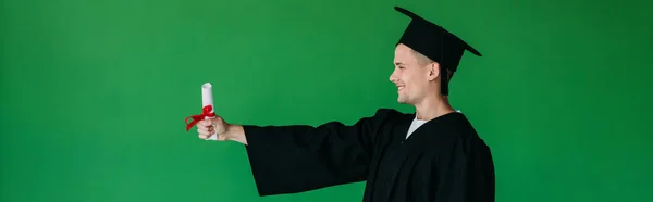 Plano panorámico de estudiante sonriente en gorra académica sosteniendo diploma aislado en verde - foto de stock