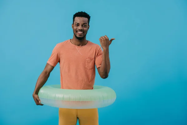 Hombre afroamericano feliz mostrando el pulgar hacia arriba y de pie con el anillo inflable aislado en azul - foto de stock