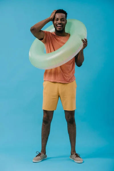 Happy african american man standing with inflatable ring and touching hair on blue — Stock Photo