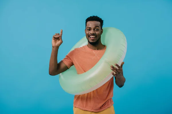 Felice uomo afroamericano in piedi con anello gonfiabile e puntando con dito isolato su blu — Foto stock