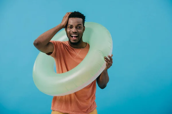 Homme afro-américain joyeux toucher les cheveux et debout avec anneau de natation isolé sur bleu — Photo de stock