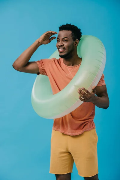 Homme afro-américain joyeux geste tout en se tenant debout avec anneau de nage isolé sur bleu — Photo de stock