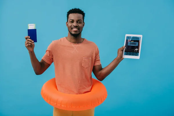 KYIV, UKRAINE - JUNE 27, 2019: cheerful african american man holding passport and digital tablet with booking app on screen isolated on blue — Stock Photo