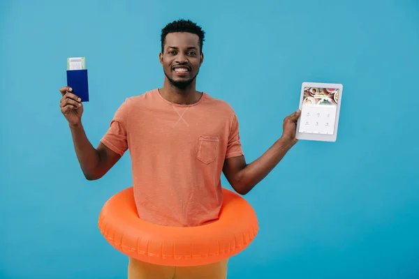 KYIV, UCRANIA - 27 de junio de 2019: alegre hombre afroamericano con pasaporte y tableta digital con aplicación cuadrada en pantalla aislada en azul - foto de stock