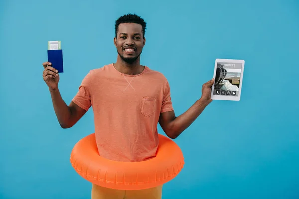 KYIV, UKRAINE - JUNE 27, 2019: cheerful african american man holding passport and digital tablet with tickets app on screen isolated on blue — Stock Photo