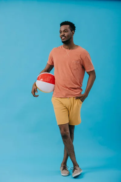 Glücklicher afrikanisch-amerikanischer Mann steht mit der Hand in der Tasche und hält Strandball auf blau — Stockfoto