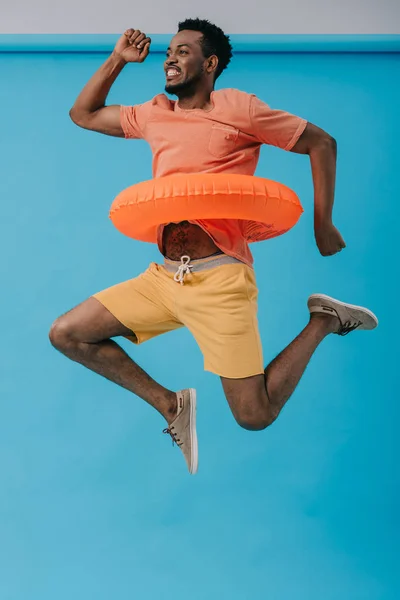 Cheerful bearded african american man jumping with swim ring on blue — Stock Photo