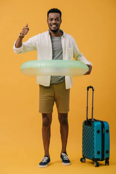 Homme afro-américain joyeux debout avec anneau de natation et bagages tout en montrant pouce vers le haut sur orange — Photo de stock