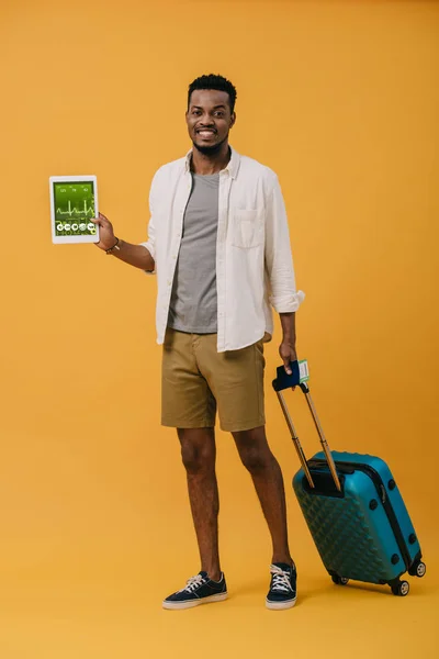 Cheerful african american man standing with luggage and holding digital tablet with medical app on screen on orange — Stock Photo