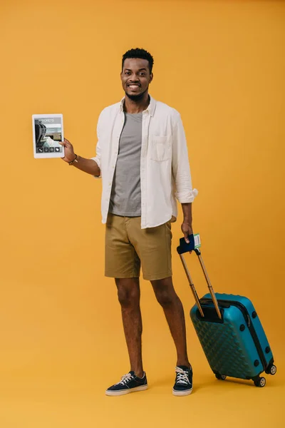 Homme afro-américain joyeux debout avec des bagages et tenant tablette numérique avec l'application de billets à l'écran sur orange — Photo de stock