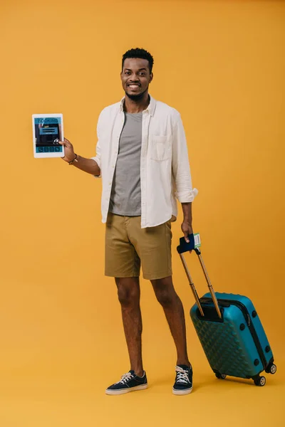 Alegre hombre afroamericano de pie con el equipaje y la celebración de la tableta digital con la aplicación de reserva en la pantalla en naranja - foto de stock