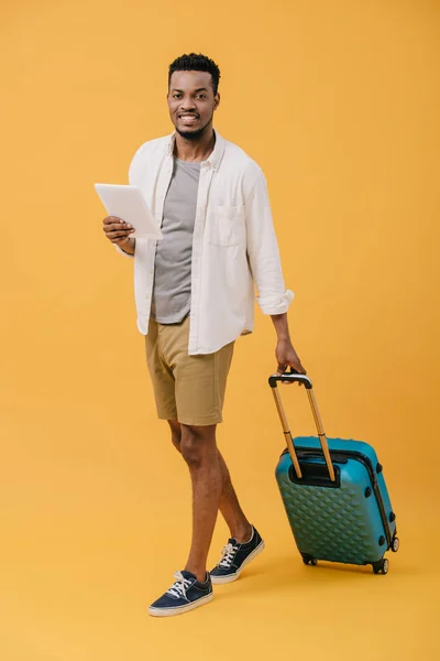 Hombre afroamericano positivo sosteniendo tableta digital y caminando con el equipaje en naranja - foto de stock