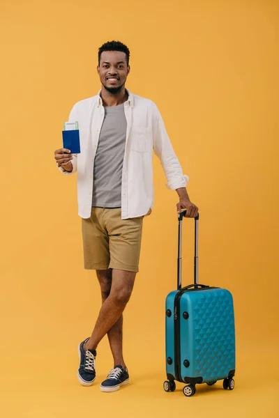 Alegre afroamericano hombre con pasaporte con billete de avión y de pie con las piernas cruzadas cerca de equipaje en naranja - foto de stock