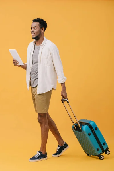 Happy african american man holding digital tablet and walking with luggage on orange — Stock Photo