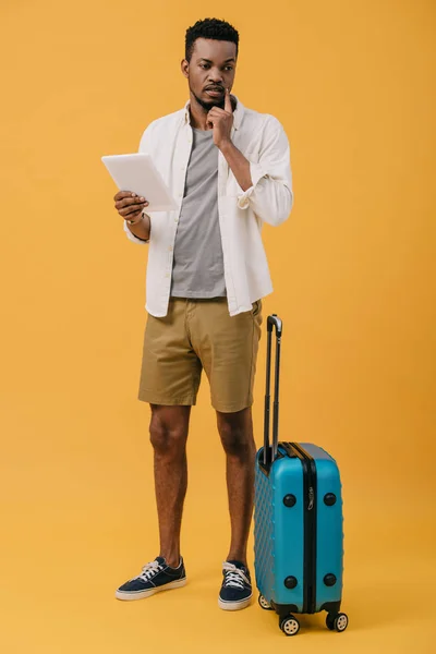 Nachdenklicher afrikanisch-amerikanischer Mann hält digitales Tablet in der Hand und steht neben Gepäck auf orange — Stockfoto