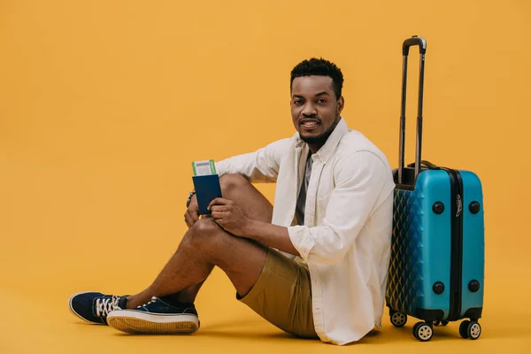 Alegre afroamericano hombre con pasaporte con billete de avión y sentado cerca de equipaje en naranja - foto de stock