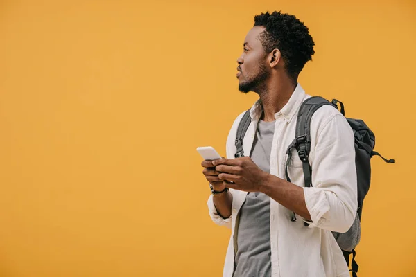 Handsome african american man with backpack holding smartphone isolated on orange — Stock Photo
