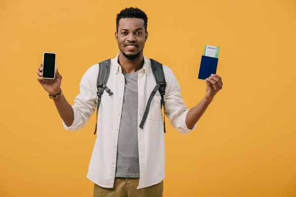 Felice uomo afroamericano con zaino in possesso di passaporto con biglietto aereo e smartphone con schermo bianco isolato su arancione — Foto stock