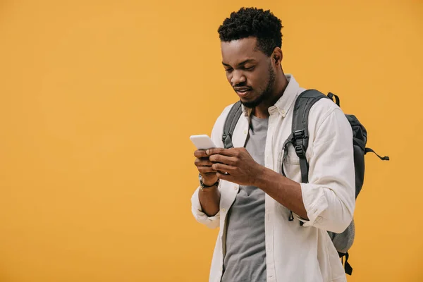 Красивий афроамериканський чоловік з рюкзаком за допомогою смартфона ізольовано на помаранчевому — стокове фото