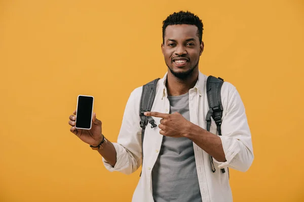 Hombre afroamericano feliz con la mochila apuntando con el dedo en el teléfono inteligente con pantalla en blanco aislado en naranja - foto de stock