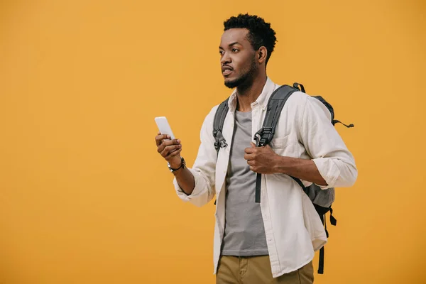 Hombre afroamericano con mochila sosteniendo teléfono inteligente aislado en naranja - foto de stock