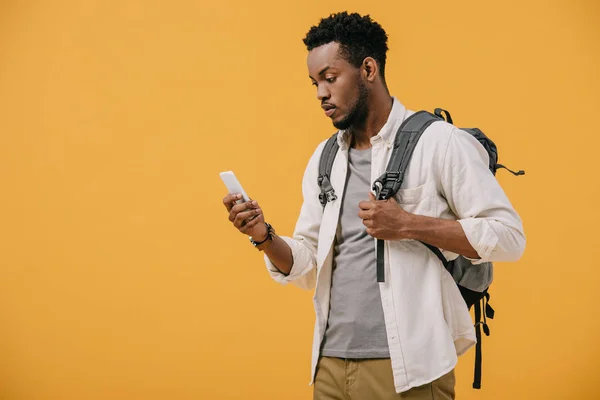 Hombre afroamericano con mochila mirando teléfono inteligente aislado en naranja - foto de stock