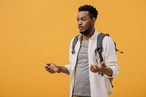 Емоційний афроамериканський чоловік з рюкзаком тримає смартфон і жестикулює ізольовано на помаранчевому — стокове фото