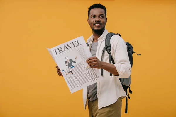 Cheerful african american man standing with backpack and holding travel newspaper isolated on orange — Stock Photo